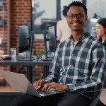 portrait-artificial-intelligence-app-developer-sitting-down-typing-laptop-fixing-glasses-looking-up-smiling-camera-programer-using-portable-computer-innovating-cloud-computing-min.jpg