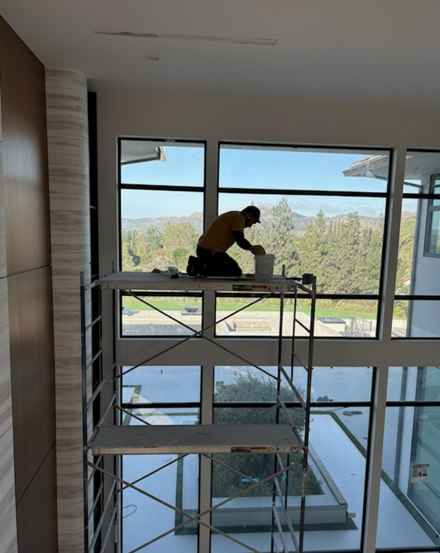 A worker on scaffolding cleans the large windows of a modern home, with a scenic view of hills and trees outside.